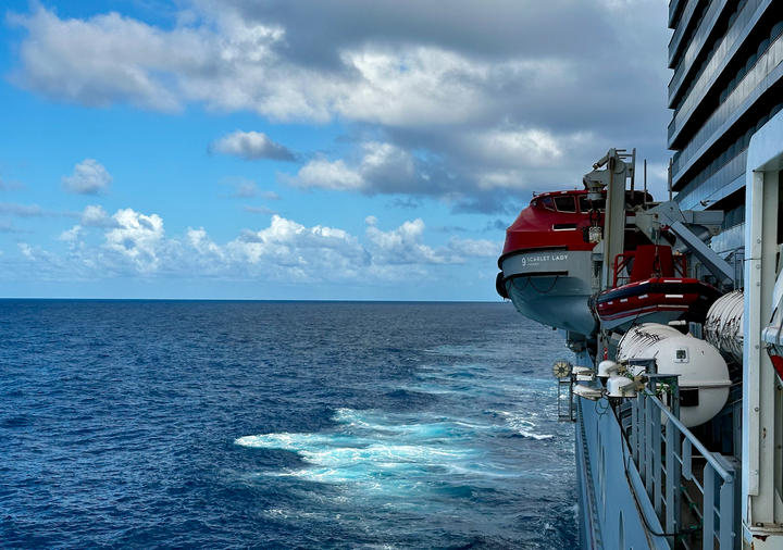 View from the side of a ship, lifeboats and railings to one side, the open ocean to the other