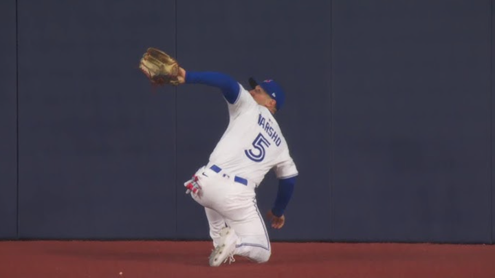 Dauton Varsho on one knee, on the warning track, his back to the field, arm out ready to make a catch