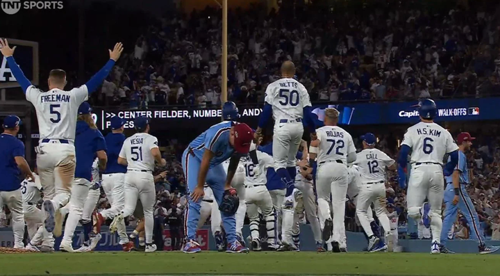 Dodgers celebrate, jumping on the field in the background while Phillies pitcher Orion Kerkering has his hands on his knees and his head hung low in the foreground