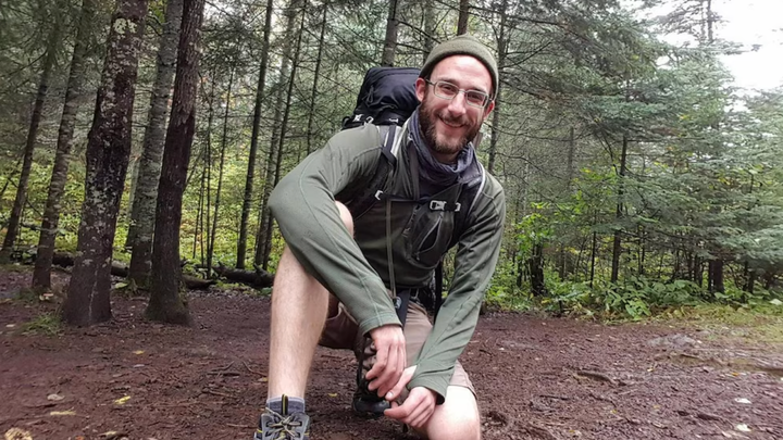 Alex Pretty in hiking gear, on one knee in the woods, smiling at the camera