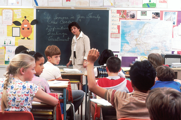 Children in a classroom looking at one student who is raising his hand.