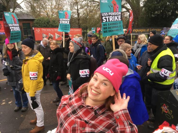 Emma wearing a pink UCU beanie and a checked shirt. She is holding her hand to her ear to hear something being said