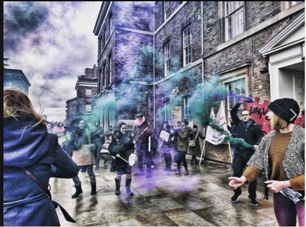 A group of strikers outside the University of Leicester on International Women’s Day (also a strike day) 2018. There are colored smoke flares and a woman beating a drum.