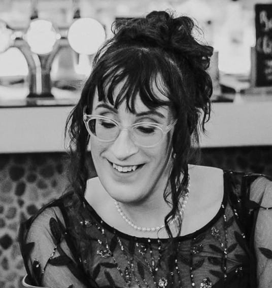 Madeleine Moore: Madeleine has long, wavy dark hair, wears glasses and is smiling as she looks down