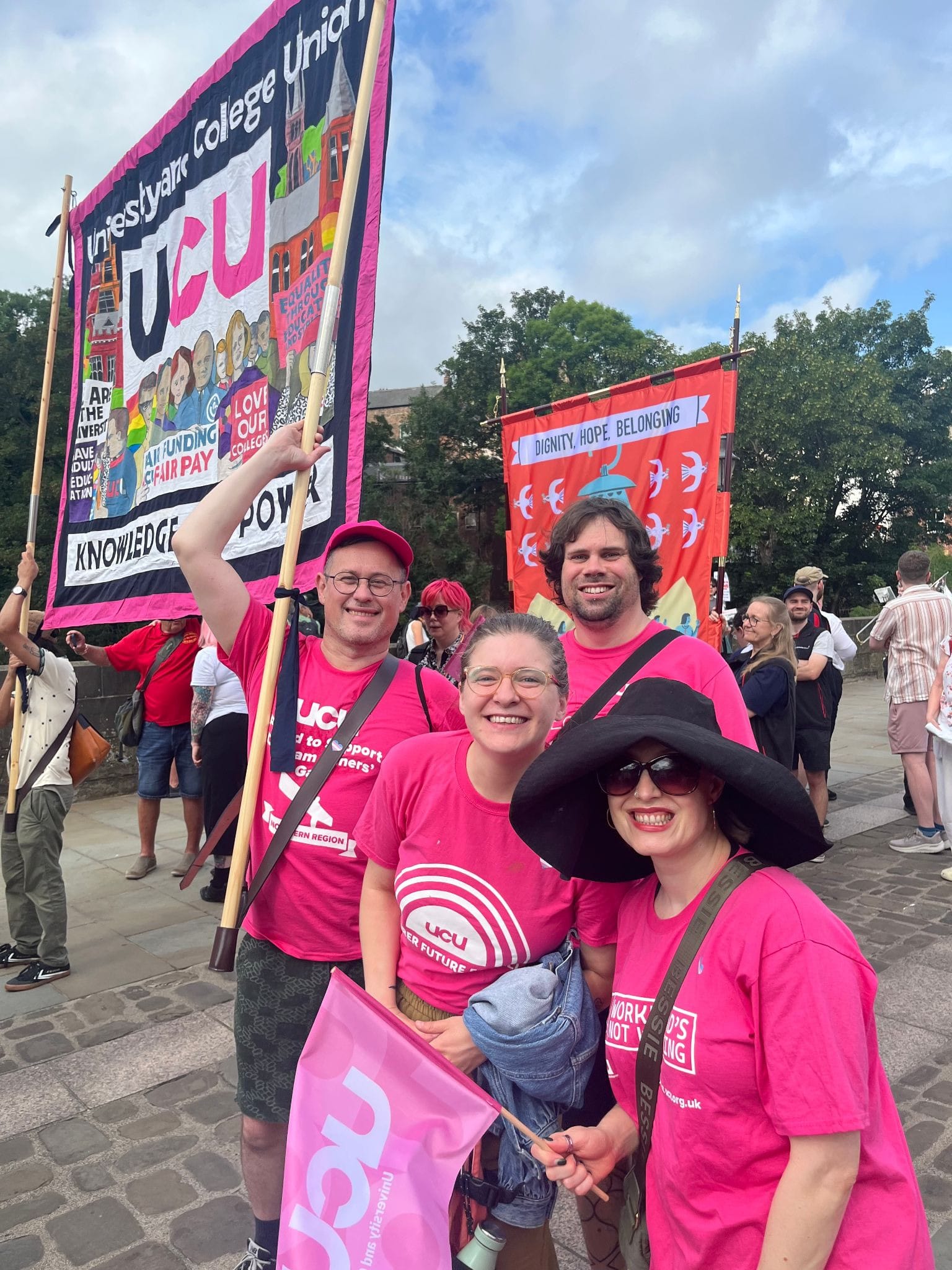 Four white people in pink UCU tshirts smiling to the camera on a cobbled road. One is holding one half of the UCU banner