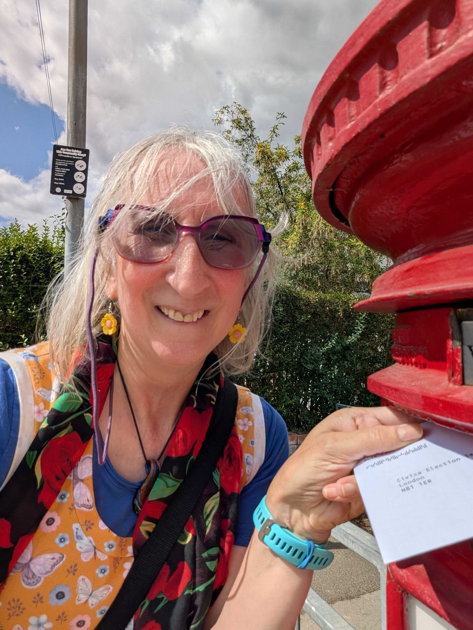A colourfully-dressed white woman smiling while posting an envelope into a red postbox in the sunshine