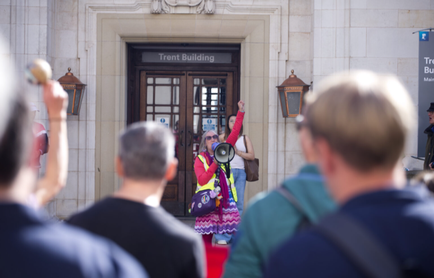 A colourfully dressed woman shouts into a megaphone in front of Nottingham's Trent Building while a crowd looks on