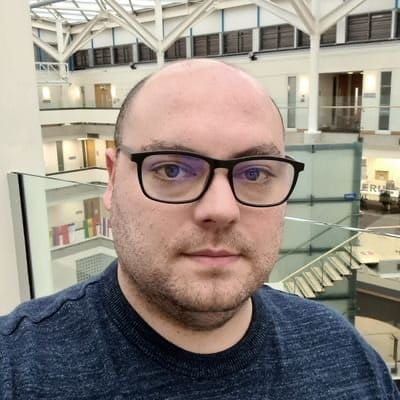 Matt Barnard - Matt is standing in an atrium. He's wearing black glasses and a dark-coloured top