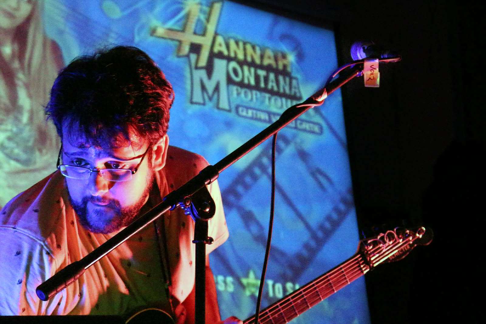 Tim holding a guitar in front of a mic stand. Projected on a large screen in the background is the menu for the video game Hannah Montana Pop Tour
