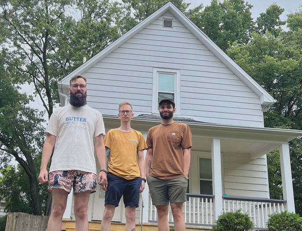 The three members of MyHeartYourGlove stand in front of a white house.