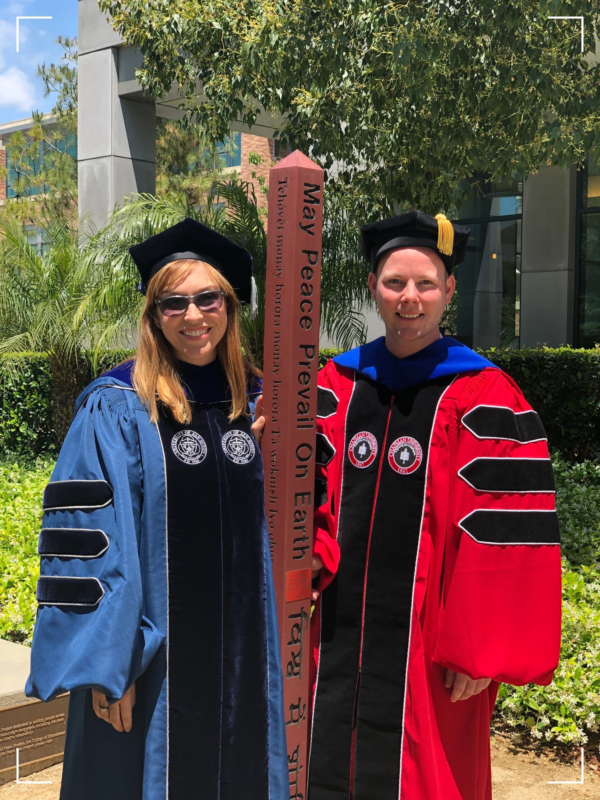 Two persons wearing doctoral regalia, one in blue and black and one in red and black, standing next to the installation of Chapman University’s Peace Pole, Orange, CA. The Peace Pole is engraged with “May Peace Prevail on Earth” in multiple languages.