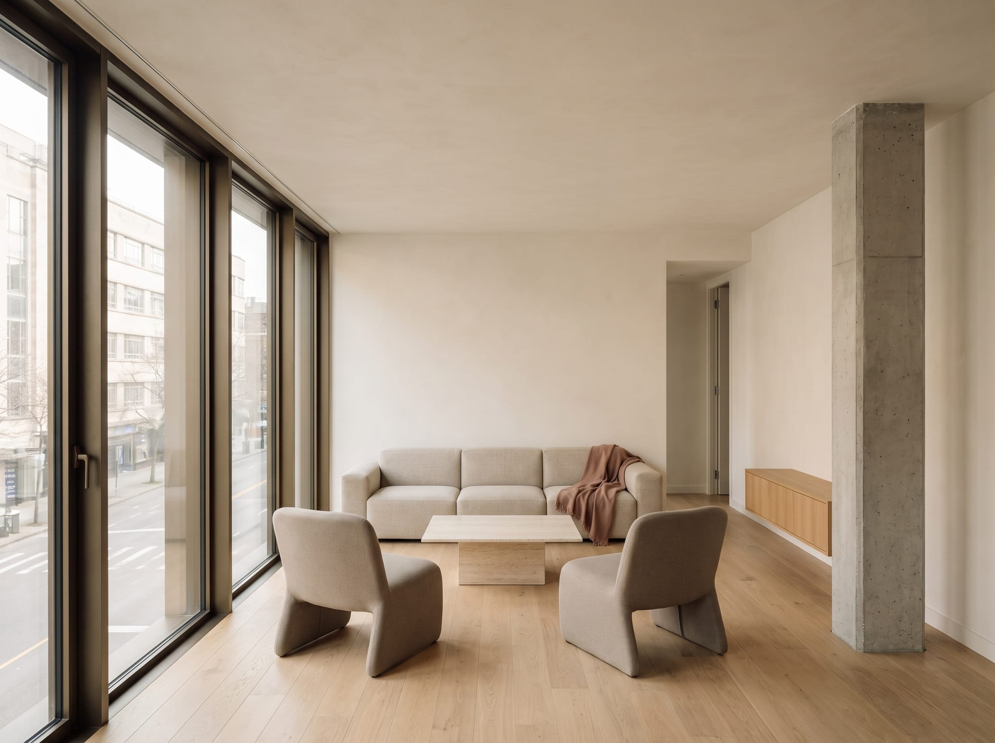 Compact apartment living room with continuous floor to ceiling glazing, one concrete column, oak flooring, and restrained furniture.