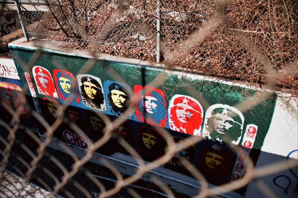 Multi-colored stencils of Che Guevara on a green background on a concrete wall surrounded by chain link fence