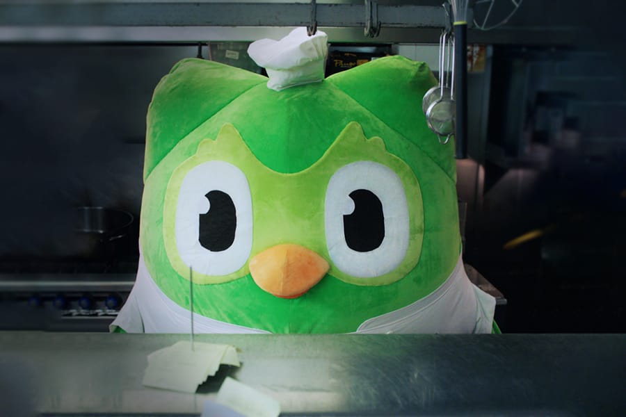 Image of Duo mascot suit wearing a chef’s hat and apron standing behind the order station in an industrial restaurant kitchen