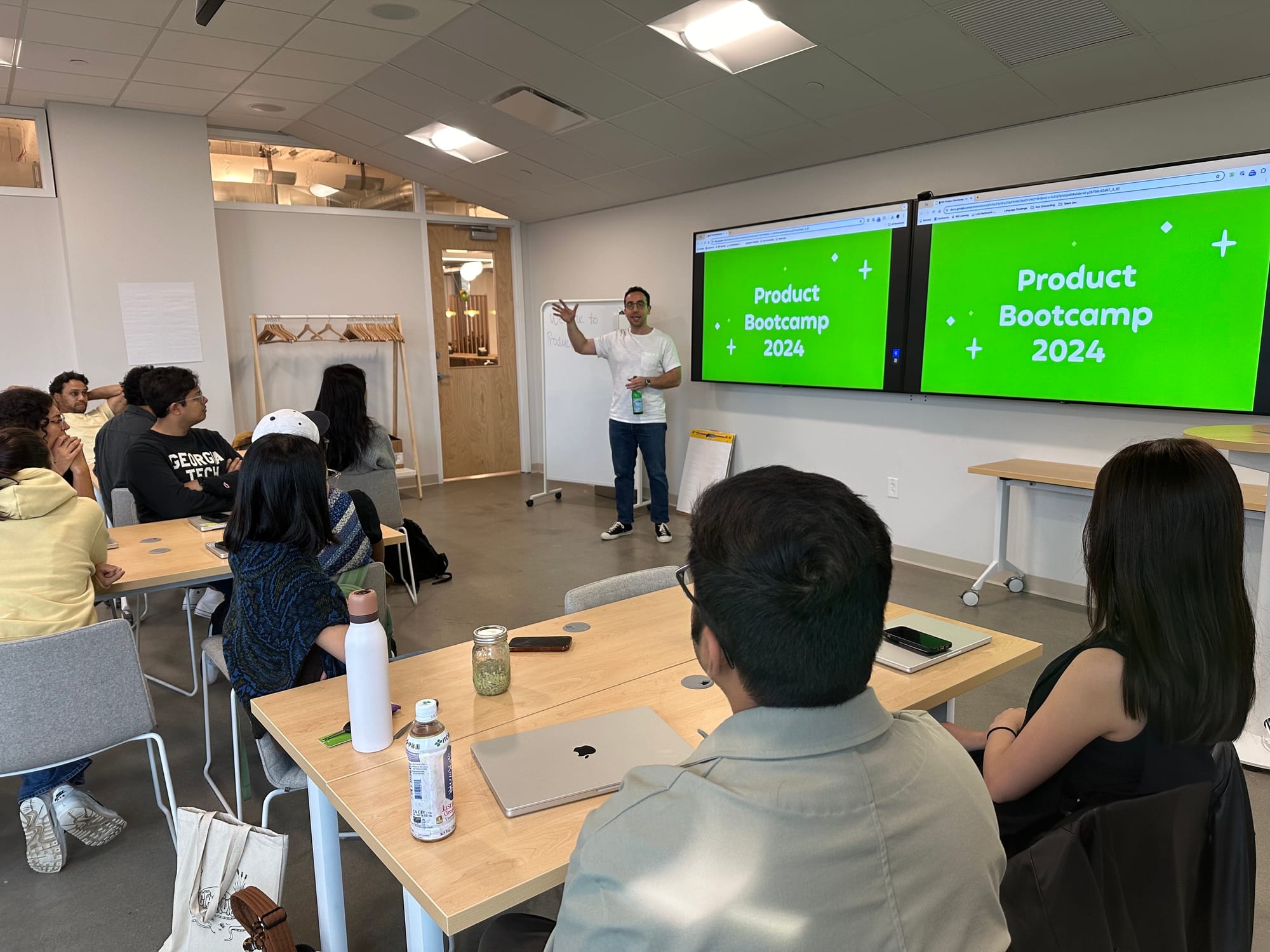 Chief Product Officer Cem Kansu presenting at Product Bootcamp 2024. He is standing at the front of a conference room with 2 screens behind him that both say Product Bootcamp 2024. We see the backs of people's heads listening to him speak.