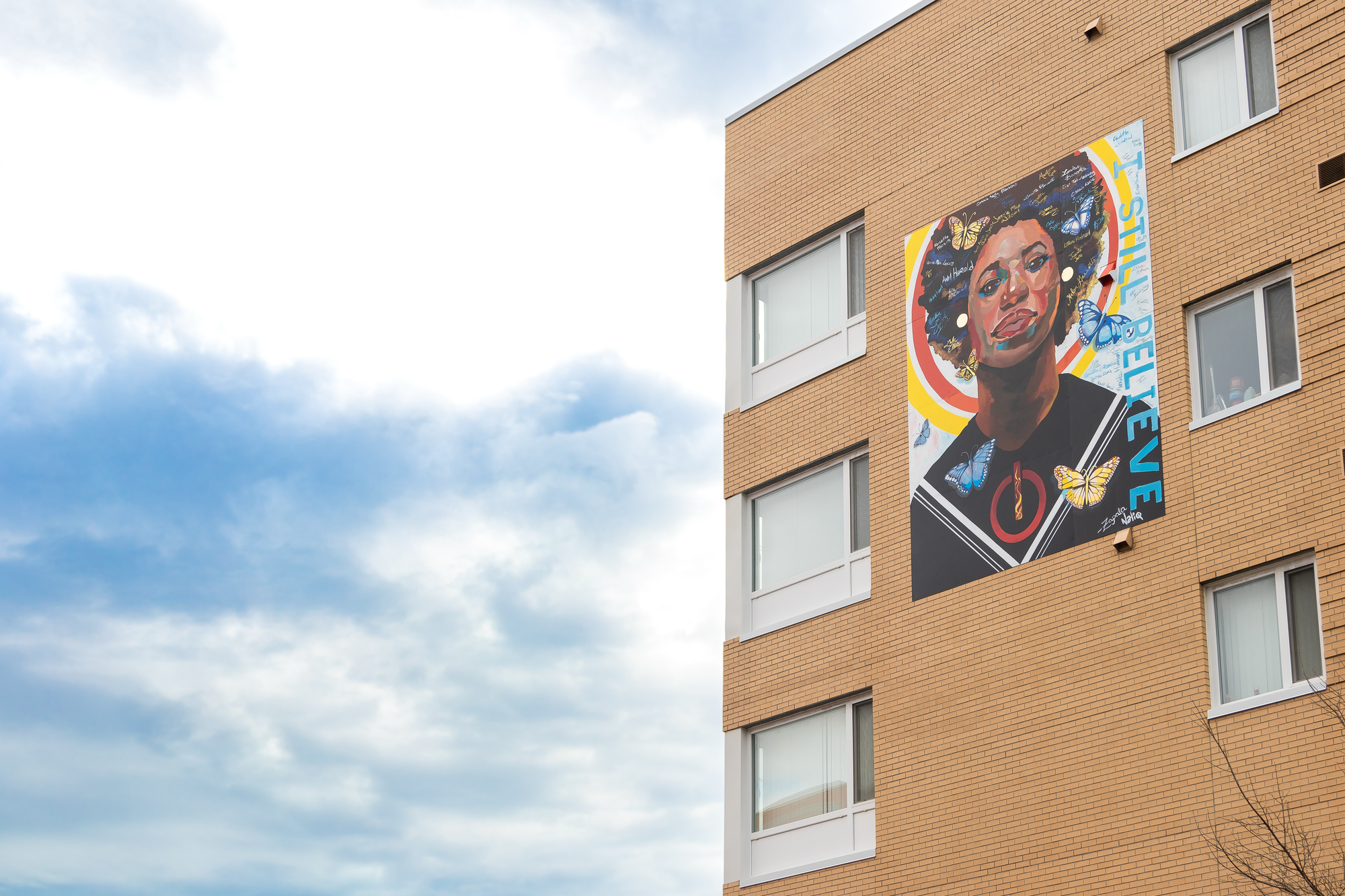 Photograph of a light-colored brick building against a cloudy and bright sky. The building features a large mural near the top floors of a Black woman with head held high and eyes looking at the street. All around her face are yellow and blue butterflies. Along the side of the mural is text that reads “I still believe.”