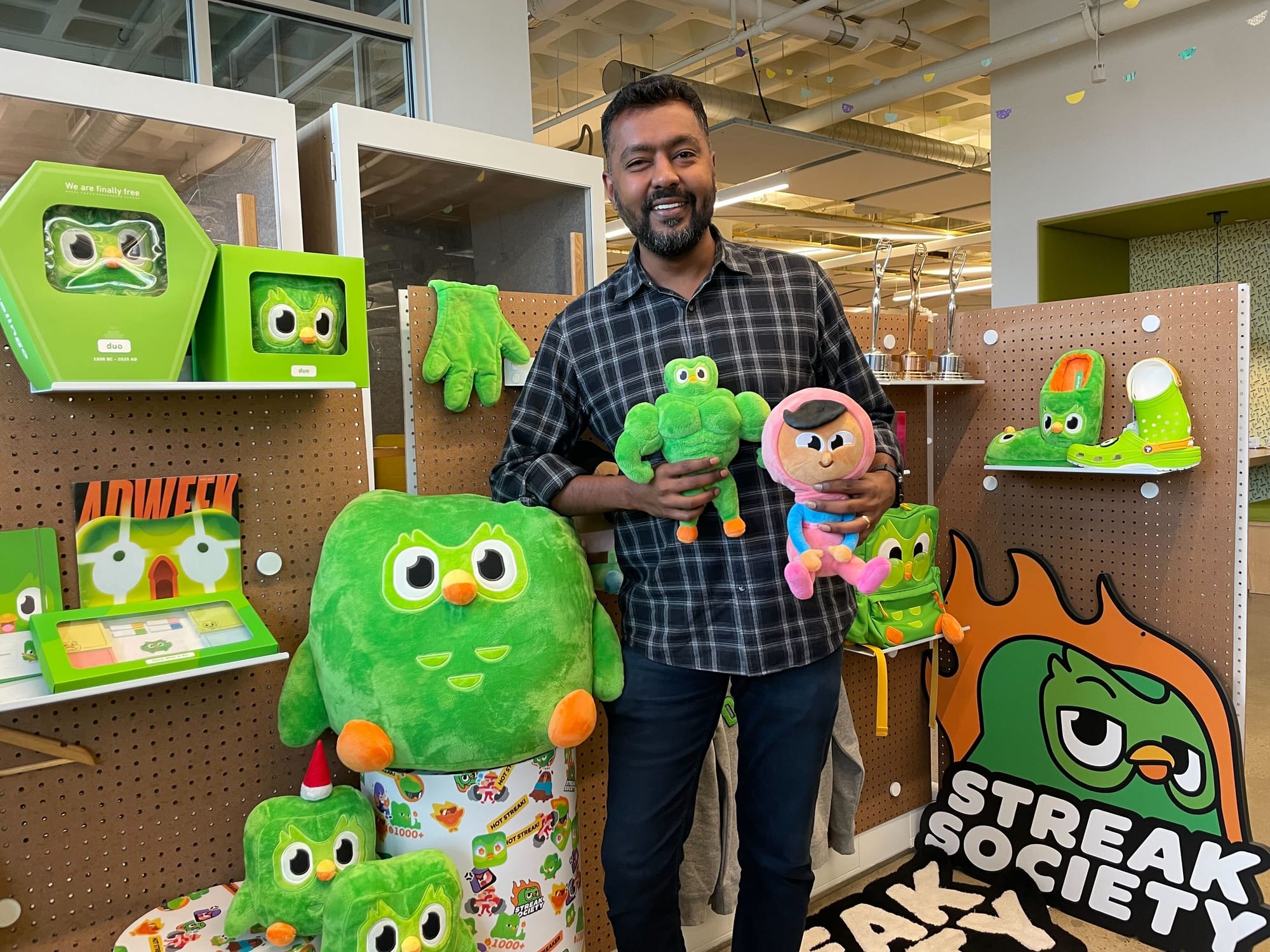Anand holding a buff Duo plushie and a Zari plushie, surrounded by various pieces of Duo merchandise. 