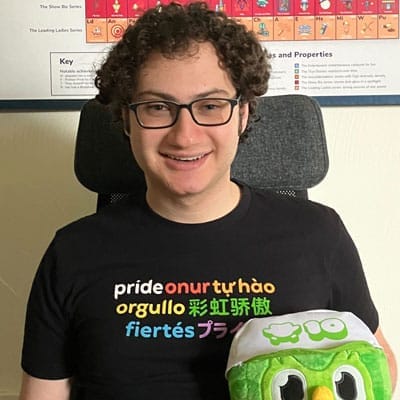 Imagen de Adam, vistiendo una camisa que dice “Pride” en múltiples idiomas y un arcoíris de colores, sentado frente a una tabla periódica de musicales de Broadway.