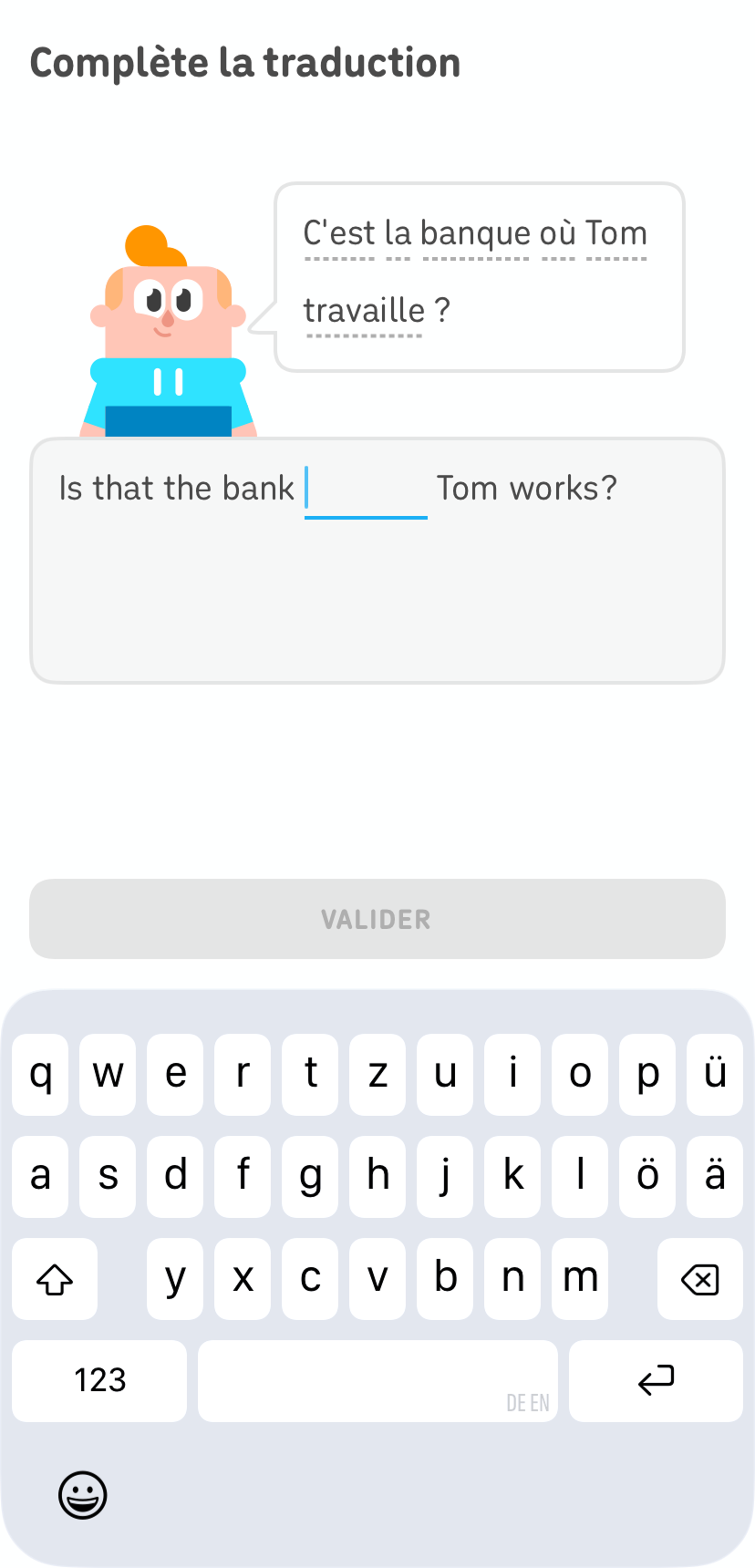 Capture d'écran d'une leçon Duolingo. La consigne indique « Complète la traduction » et Junior, l'un des personnages de Duolingo, se tient debout à côté d'une bulle de texte qui affiche la phrase en français « C'est la banque où Tom travaille ? » En dessous, on peut voir une phrase partiellement remplie en anglais avec un mot manquant.
