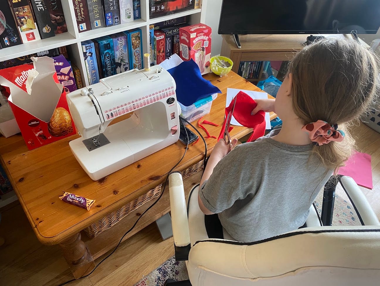 A little girl sits at a coffee table, sewing two pieces of felt together using a sewing machine. Also on the table, there are Easter eggs