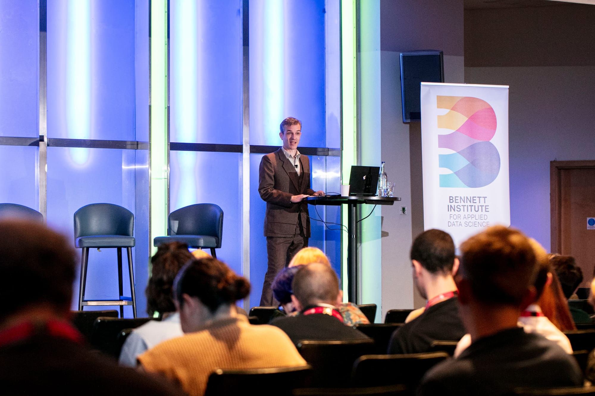 Seb Bacon stands on stage at the OpenSAFELY Community Symposium, giving a presentation