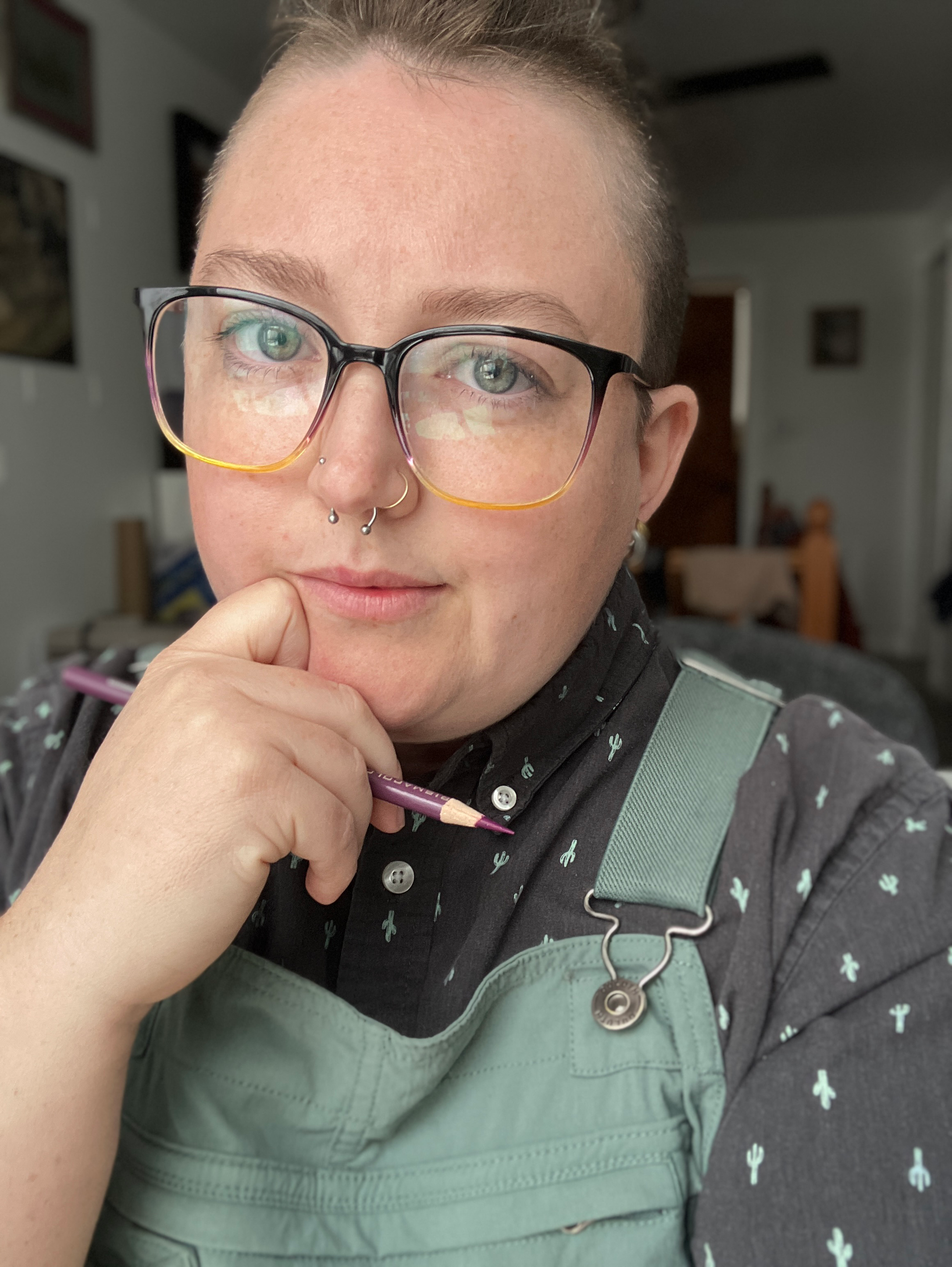 A white person with brownish greyish hair, freckles, and greyish greenish eyes is wearing glasses, a cactus button-up shirt, and overalls, and holding a pencil.