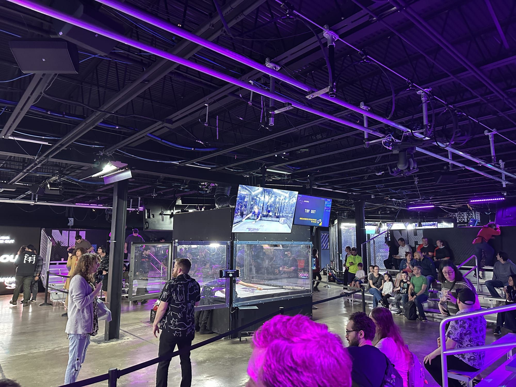 NHRL combat arena. Shot from stands. Hosts on microphone bottom left. Cage 2 center with screens between fight. Additional cages in back. Lights and cameras visible on top.