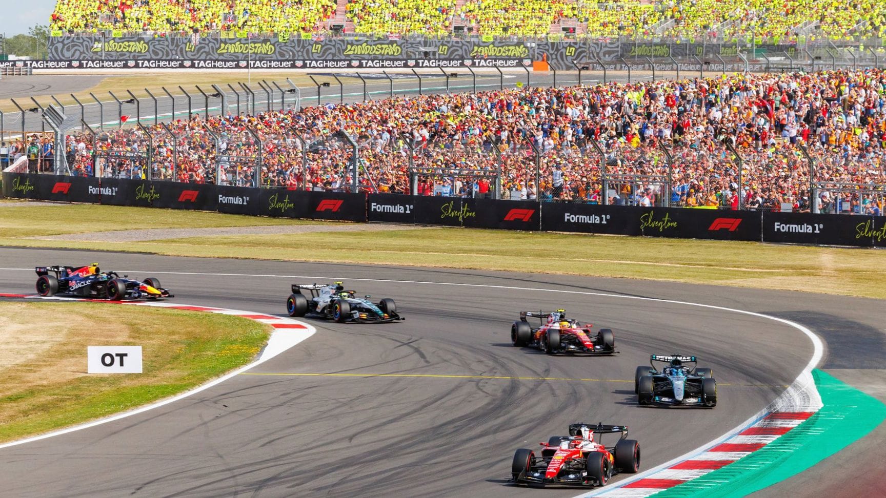 Formula 1 cars race through a corner at Silverstone in front of grandstand spectators.