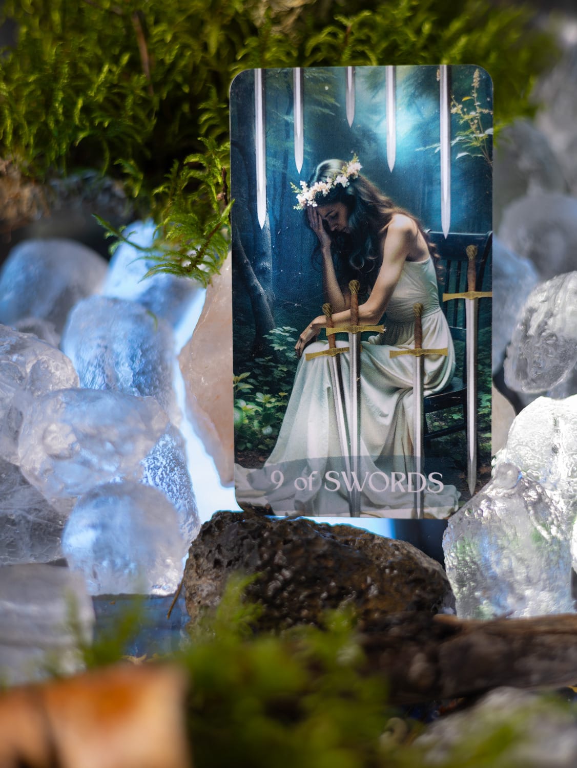 Nine of Swords tarot card framed by clear crystals, moss and cool blue light.