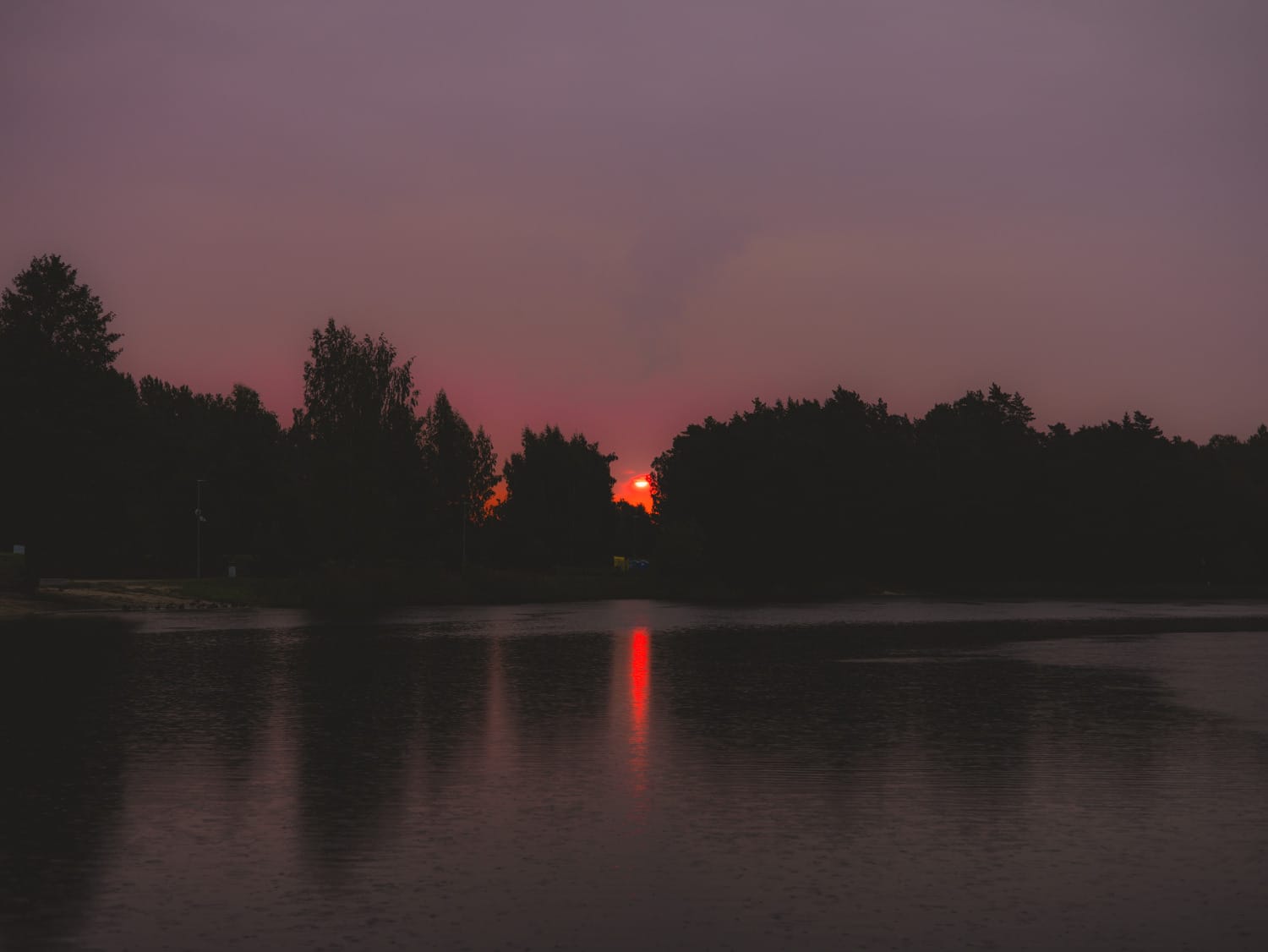 Deep red sunset disappearing behind a dark tree line, reflected vertically across calm water.