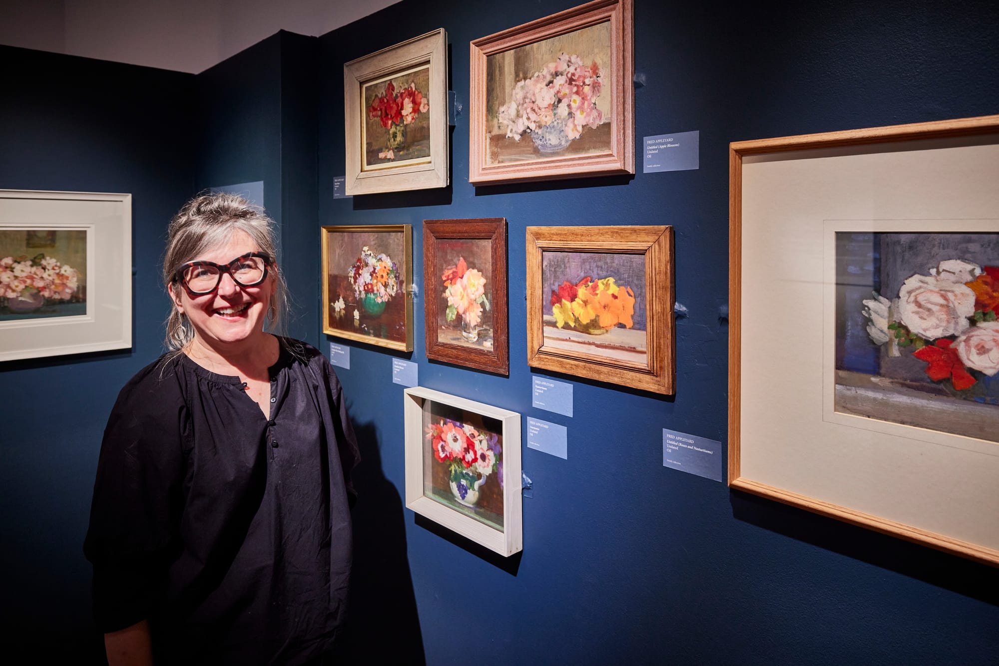A woman standing next to Fred's paintings in the exhibition, smiling