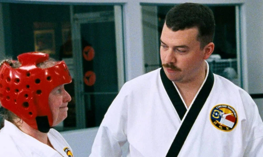 Danny McBride as Fred Simmons in The Foot Fist Way. He has a moustache, is wearing a martial arts uniform, and is speaking to a woman in a red sparring helmet.