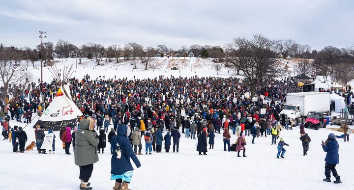 Hundreds of people in winter coats and boots stand on a snowy field. In the background are bare trees and a cloudy sky.