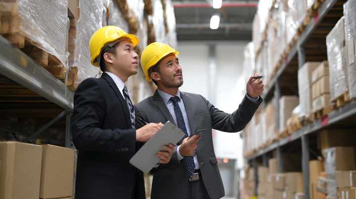 두 명의 남성이 정장을 입고 노란 안전모를 착용한 채 창고 선반을 점검하고 있다. 한 사람은 태블릿에 기록하고, 다른 사람은 손으로 선반 쪽을 가리키며 설명하고 있다. 주변에는 포장된 박스들이 높게 쌓여 있다