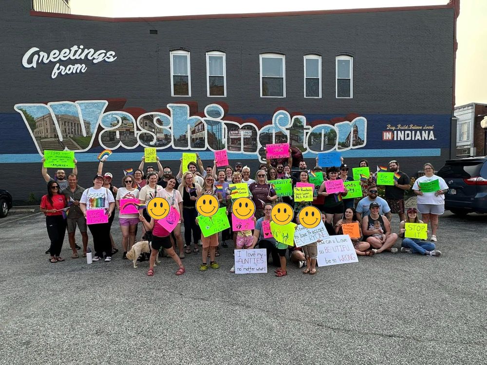 A group of pro-LGBTQIA+ protesters hold up neon colored signs with positive messages in front of a large grey brick building with a mural that says "Greetings from Washington, Indiana"