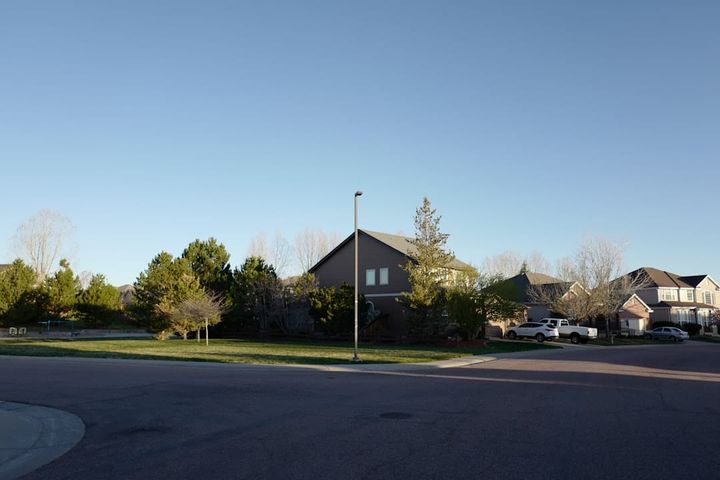A two-story house set behind trees across a wide residential street under a clear sky