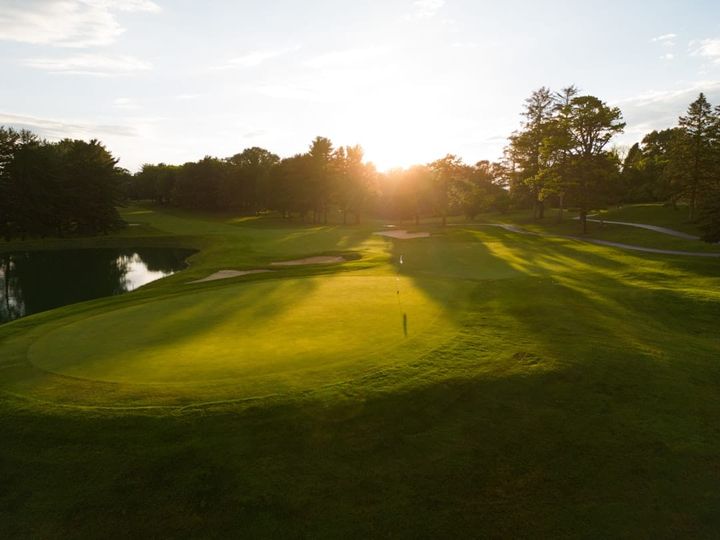 A manicured golf green with a flagstick, sand bunkers, and a small water hazard ringed by mature trees, lit low by the sun near the horizon.