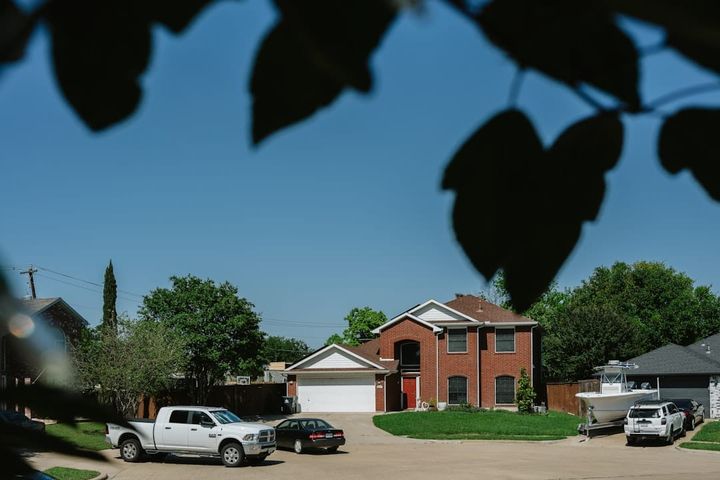 A two-story brick single-family home with a two-car garage, a pickup and a car in the driveway and a boat on a trailer alongside, framed by green trees under a clear blue sky.