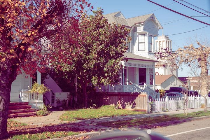 An older Victorian house with a front porch and a white picket fence on a tree-lined residential street in autumn.