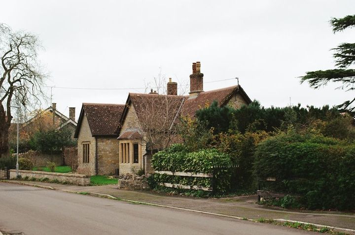 A modest single-story older cottage with a tiled roof and brick chimneys, behind a low stone wall and hedges on a quiet, empty street under an overcast sky.