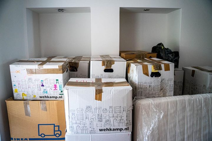 A room full of stacked, taped cardboard moving boxes and a plastic-wrapped mattress against bare white walls, a home mid-move.
