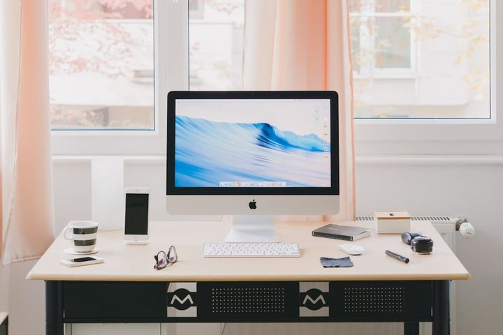 A tidy home-office desk with an all-in-one computer, keyboard, phone, and coffee mug by a bright curtained window