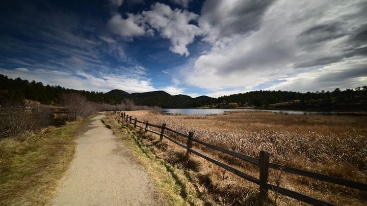 A dirt trail runs beside a wooden split-rail fence past golden marsh grass to a lake, with forested foothills and dramatic clouds behind.