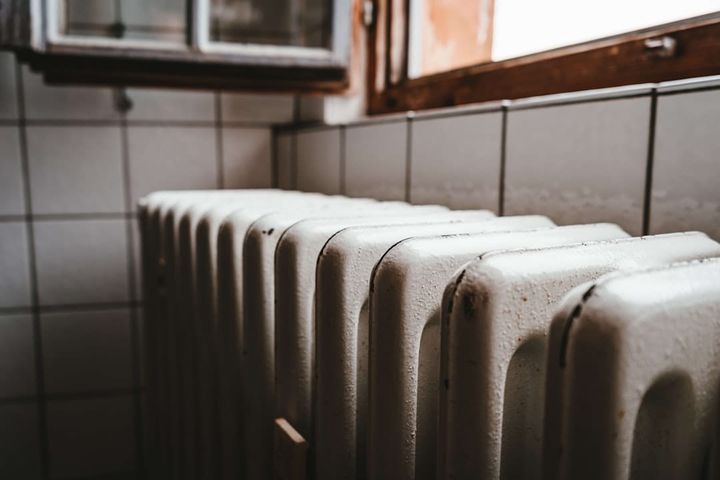 An aging cast-iron radiator with chipped, rust-flecked paint below a window whose wooden frame is weathered and peeling, against an old tiled wall.