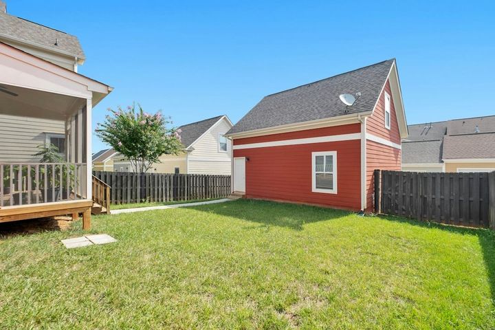 A detached two-story red outbuilding with a side door and an upstairs window at the back of a fenced lawn, behind a house with a screened porch.