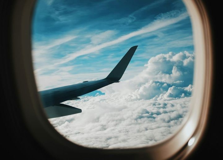 A view through an aircraft cabin window: the wing in silhouette against blue sky, with a thick bank of cloud below. No people are in frame.