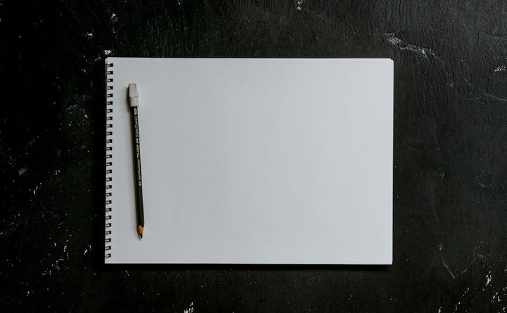 A blank spiral-bound pad lying open on a dark scuffed surface in flat overhead light, a pencil resting against its left edge, nothing written on the page.