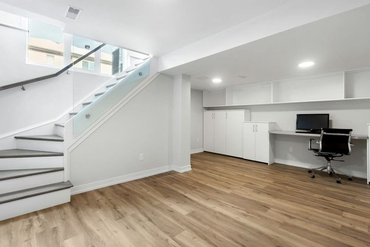 A finished basement with a stairway rising beside high windows, light wood-look flooring, white built-in cabinets and a desk with an office chair