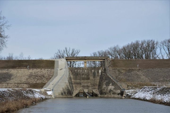 A concrete water-control structure spanning a canal on flat open ground, with bare trees on the far bank and snow along the edges.