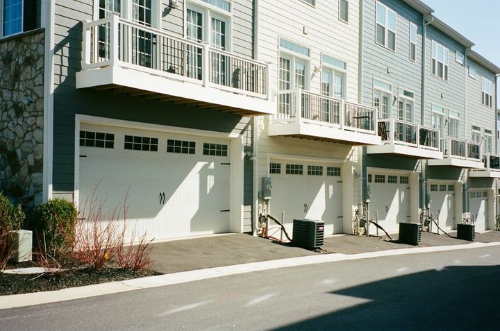 A row of modern attached townhomes in daylight, each with a white garage door below a balcony, and air-conditioning units along the shared drive.